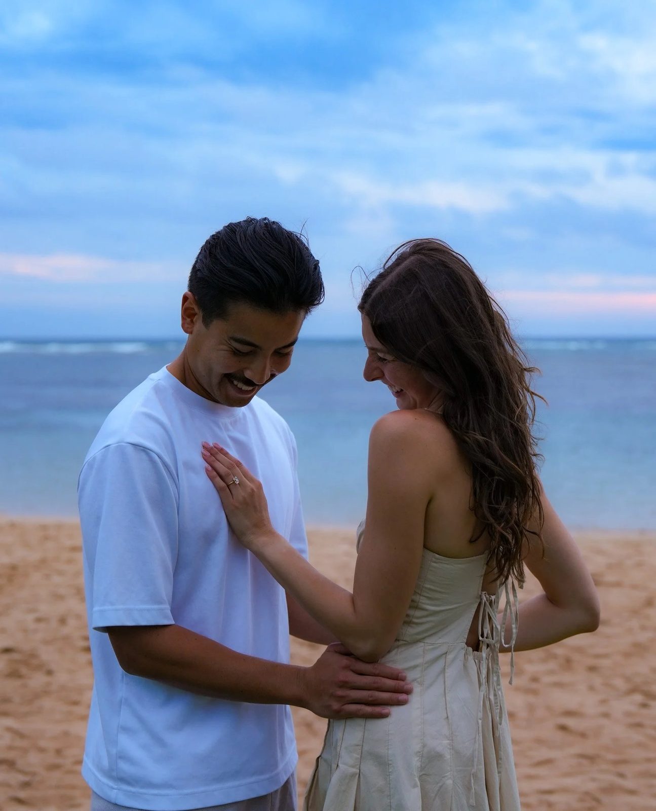 A couple laughing together on the beach, her hand with the new ring resting on his chest