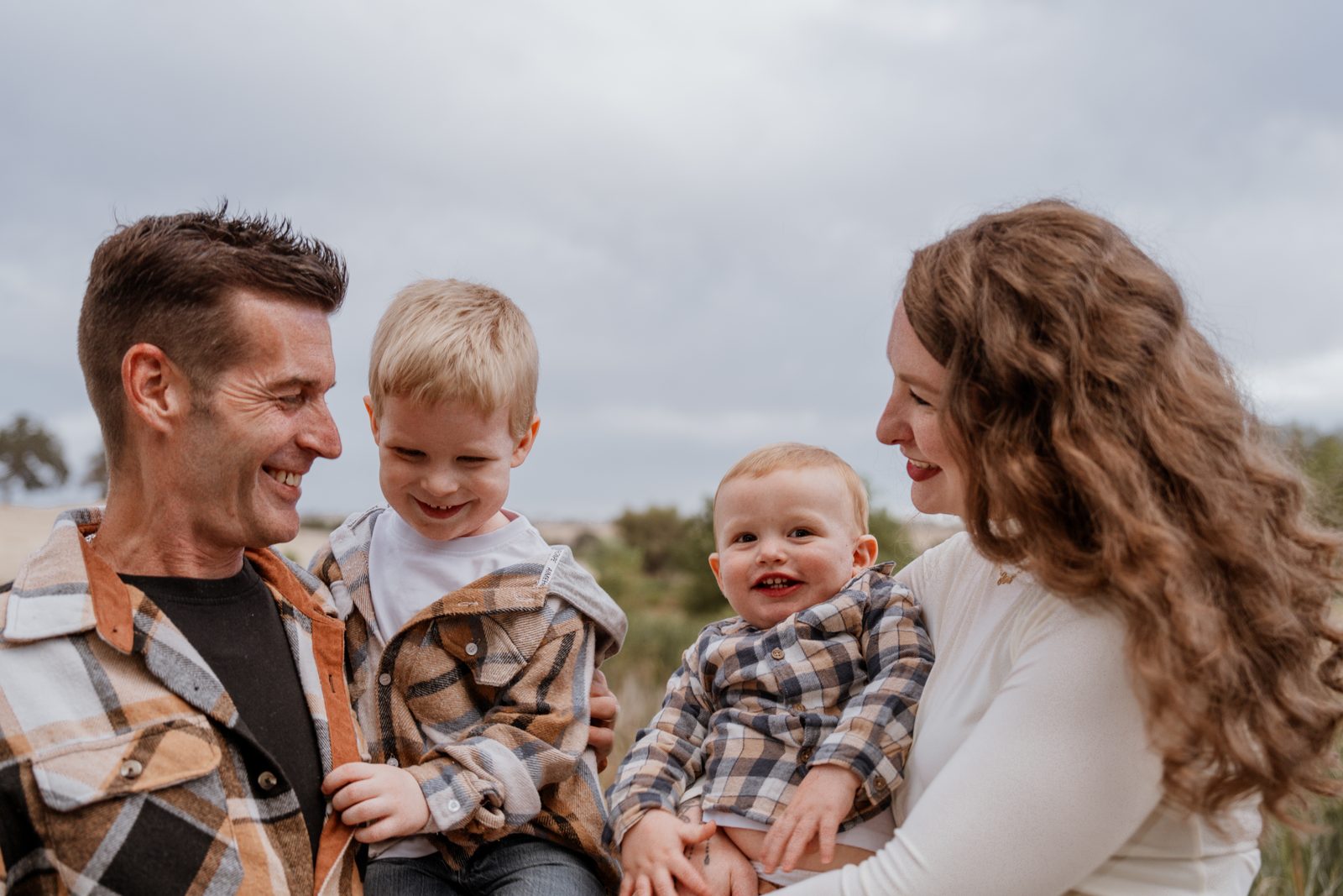 A family of four laughing together, the father grinning at his older son and the mother smiling at the baby on her hip