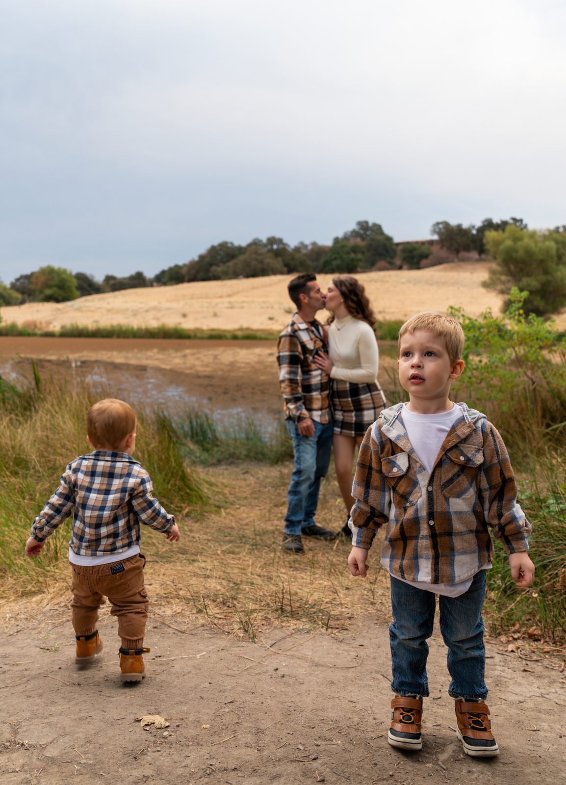 The older boy stands in the foreground looking up and away while the toddler walks off toward the long grass and the parents kiss behind them beside the pond