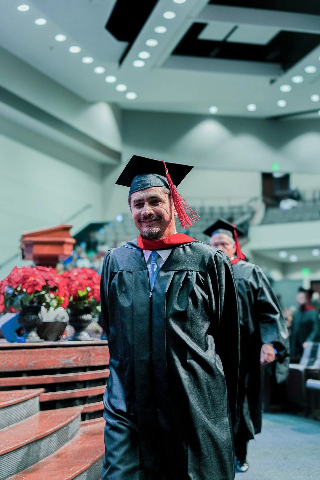 A graduate in cap and gown walks down the aisle with a small, proud smile, his red tassel swinging