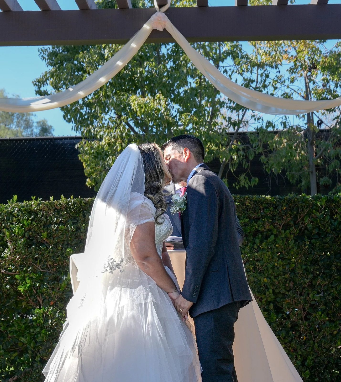 A bride and groom kiss beneath the draped pergola at their ceremony, sunlight glowing through her veil