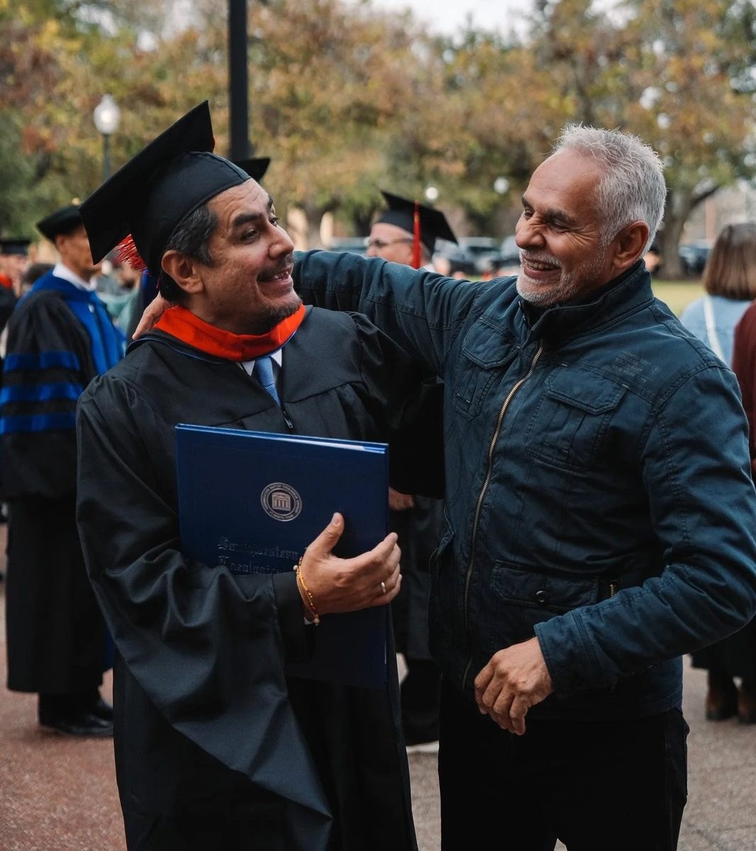 A graduate in cap and gown laughing as an older man pulls him into a one-armed hug